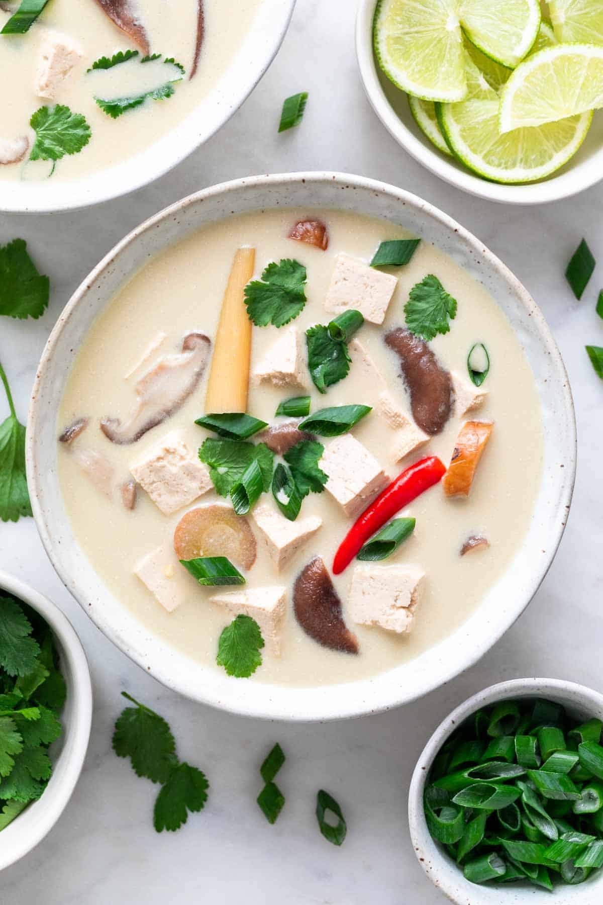 Two bowls of vegan tom kha soup with lime, scallions, and cilantro around.