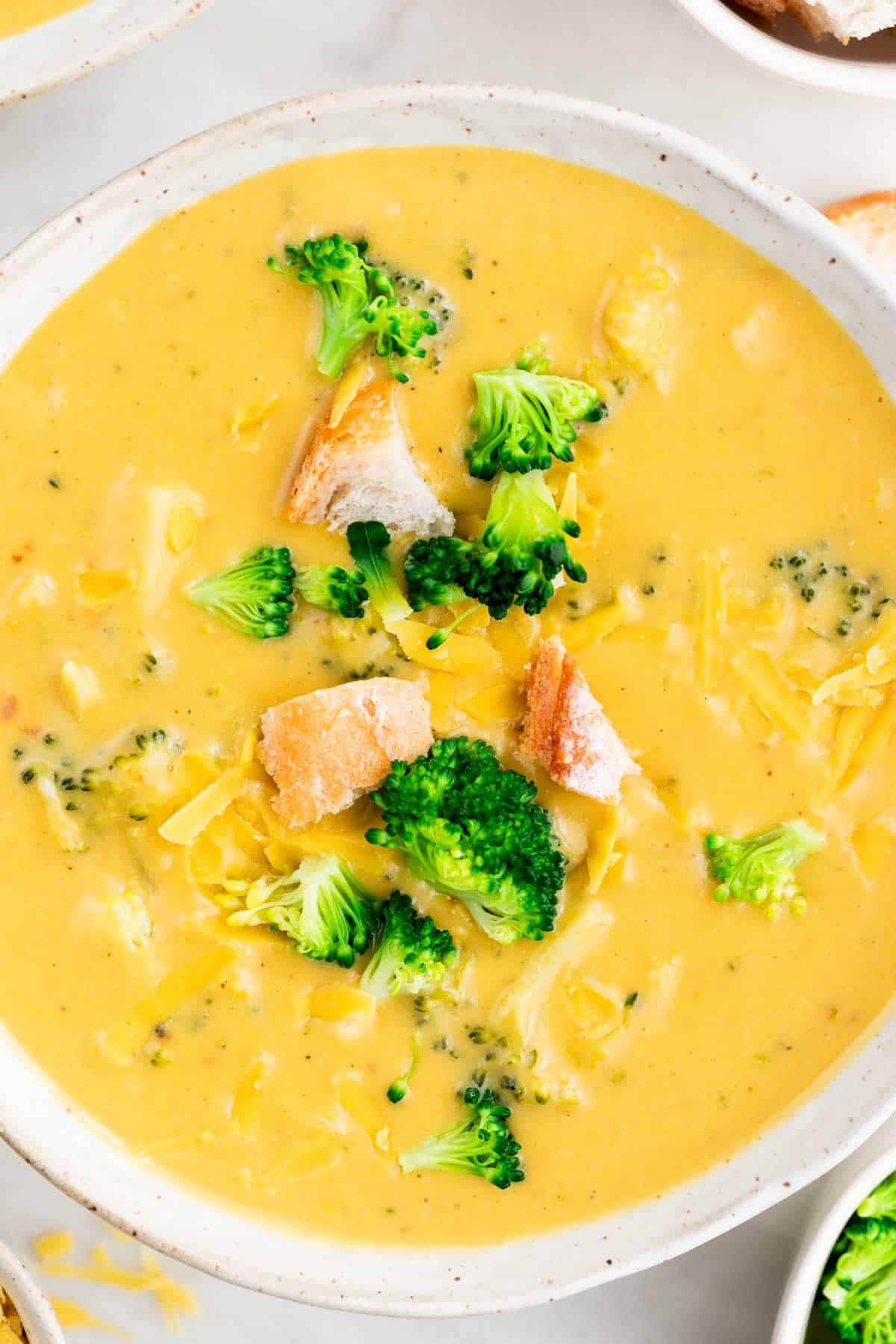 Vegan broccoli cheddar soup in a bowl topped with bread and cheese, surrounded by additional toppings.