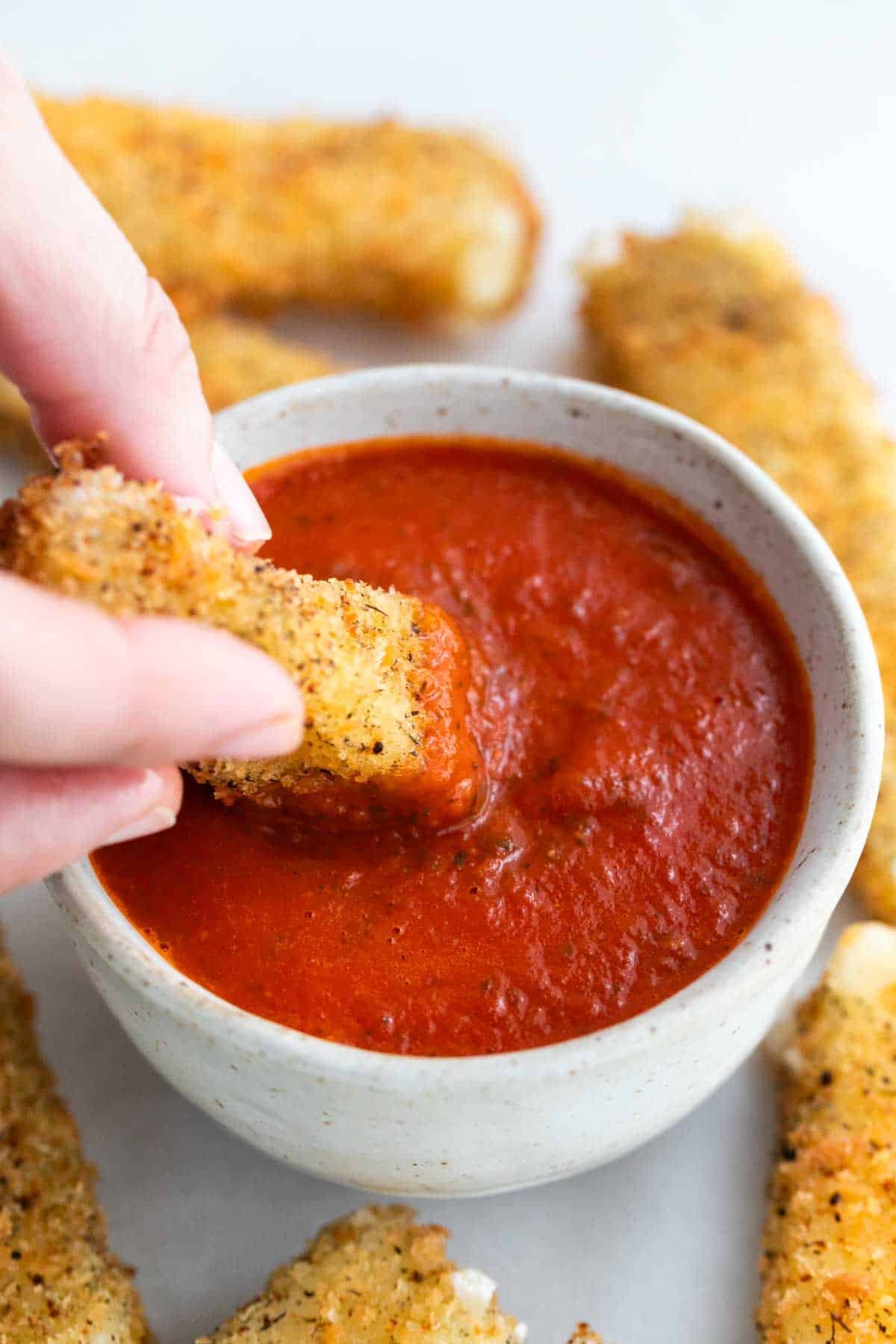 A hand dipping a vegan mozzarella stick into marinara sauce.