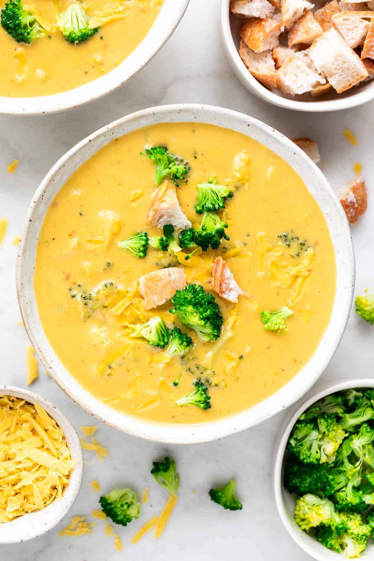 Vegan broccoli cheddar soup in a bowl garnished with bread and cheddar cheese, with toppings around.