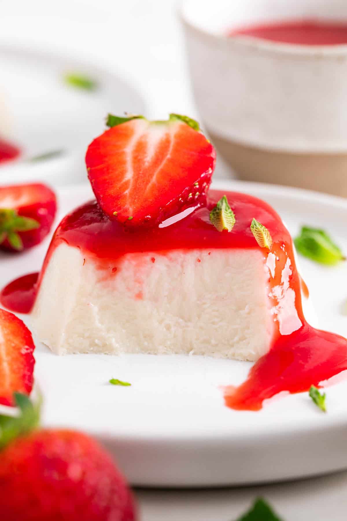 Sliced vegan panna cotta on a plate, showing the inside, with strawberry coulis, a strawberry on top, and mint leaves.