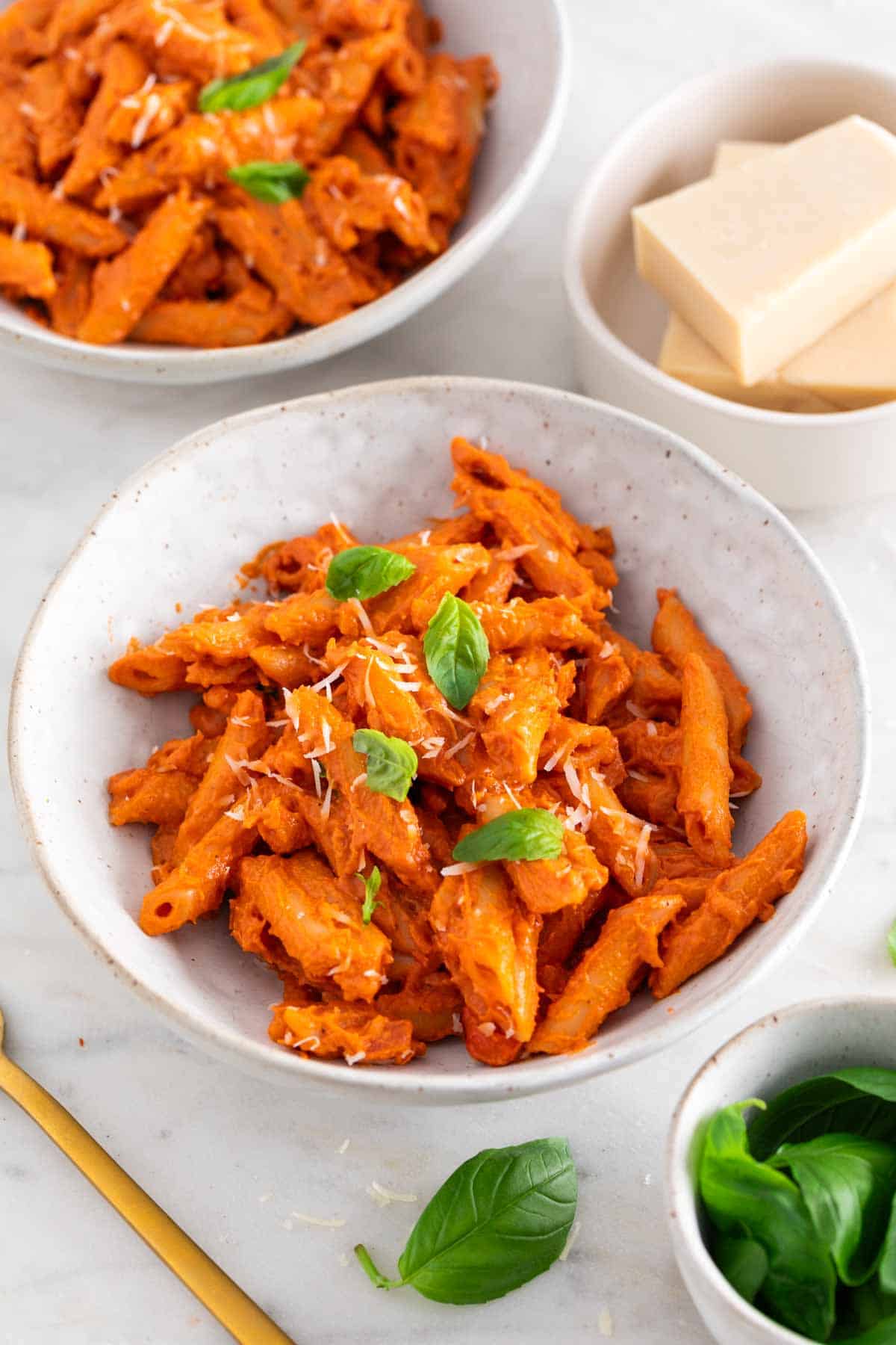Two bowls of vegan penne alla vodka, surrounded by basil and vegan parmesan cheese.