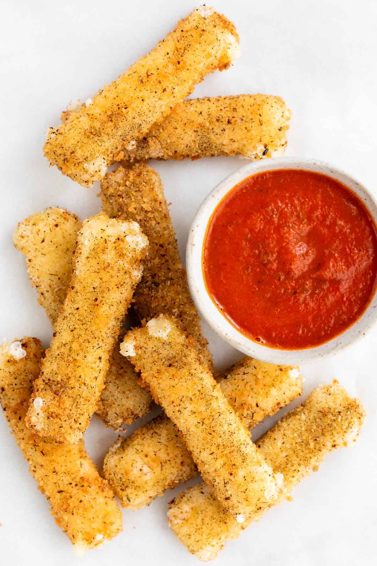 Vegan mozzarella sticks on a white background with a small bowl of marinara sauce.