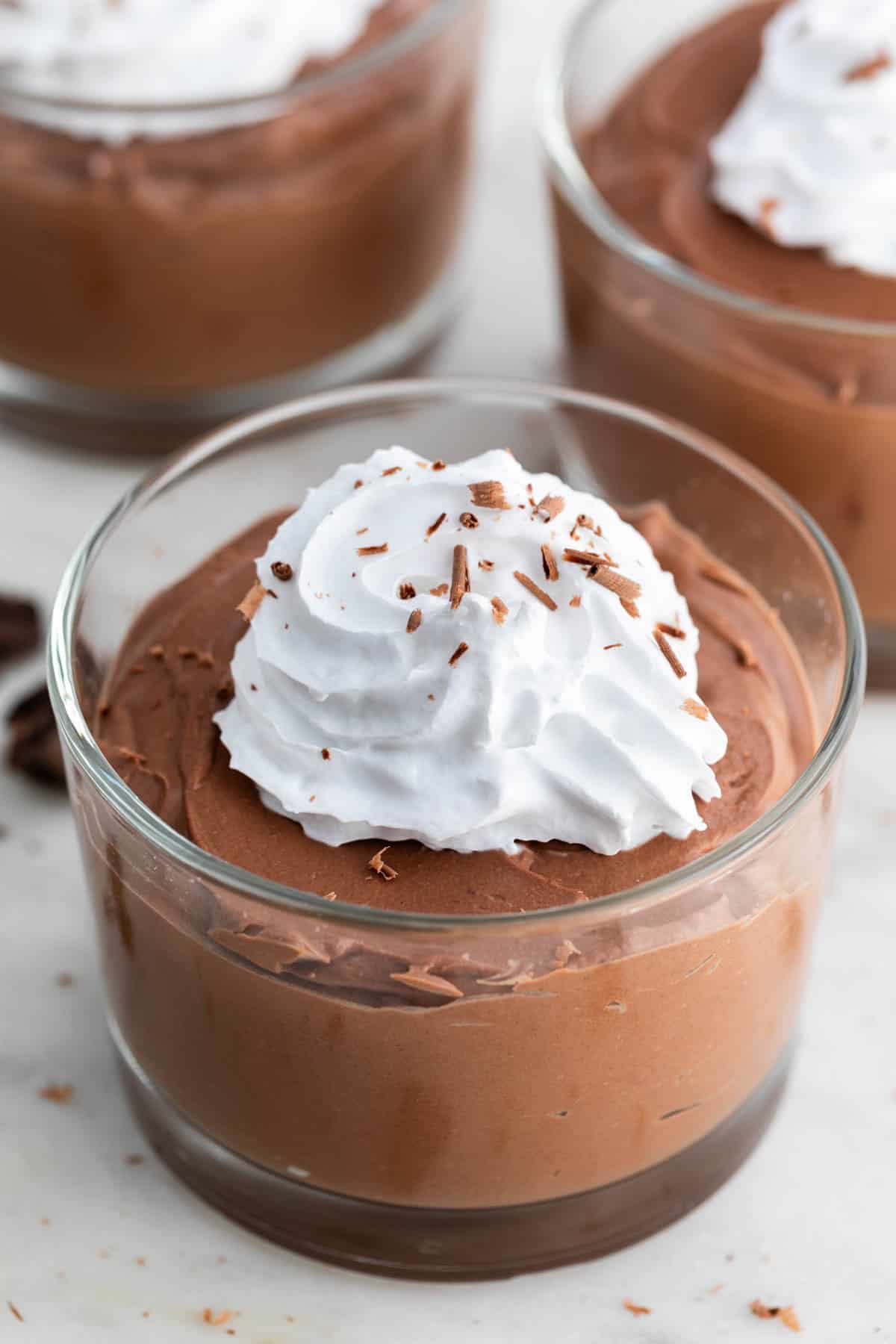 Three serving glasses of tofu chocolate mousse topped with vegan whipped cream and chocolate shavings.