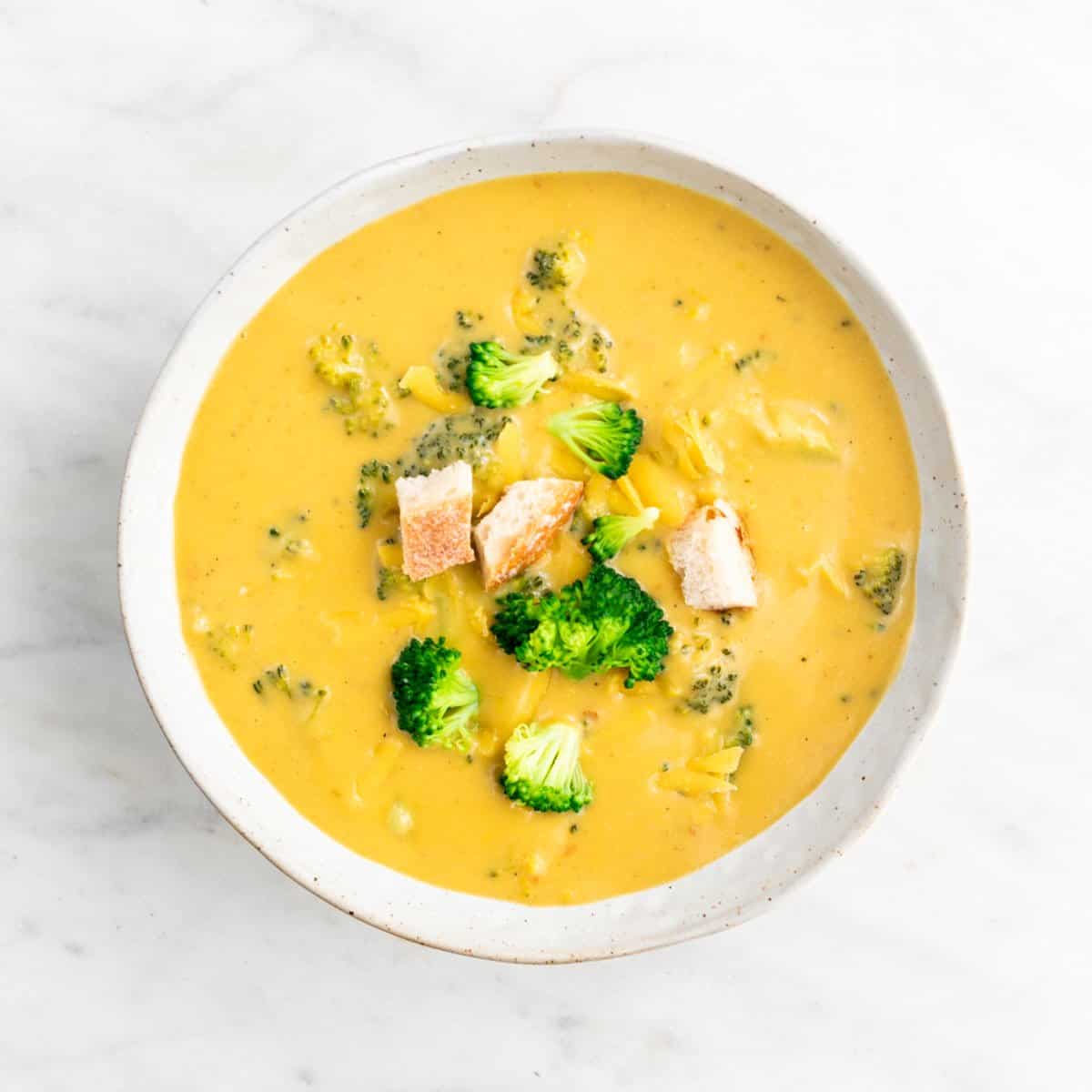 Vegan broccoli cheddar soup in a bowl garnished with bread and vegan cheddar cheese.