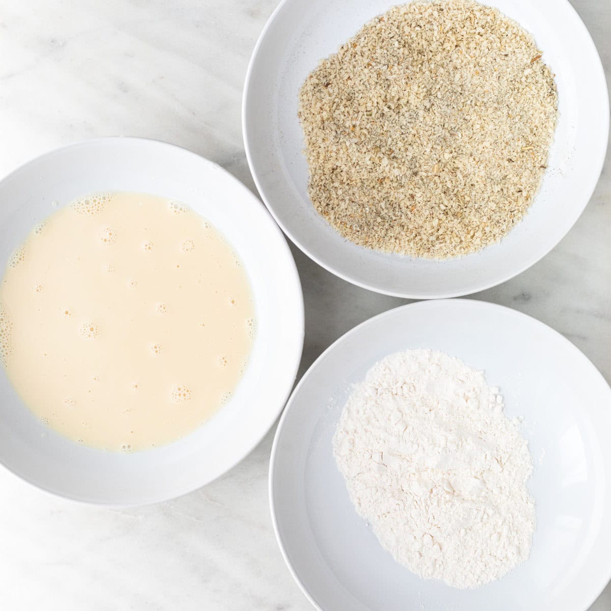 Three shallow dishes prepared for coating vegan mozzarella sticks.