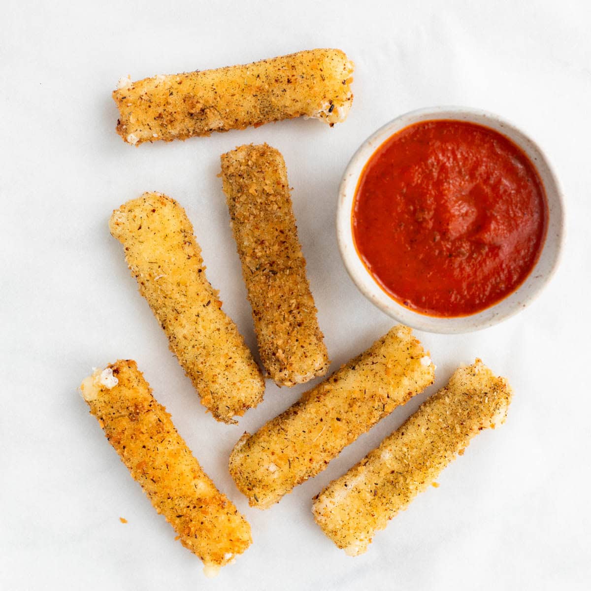 Vegan mozzarella sticks on a white background with a small bowl of marinara sauce.