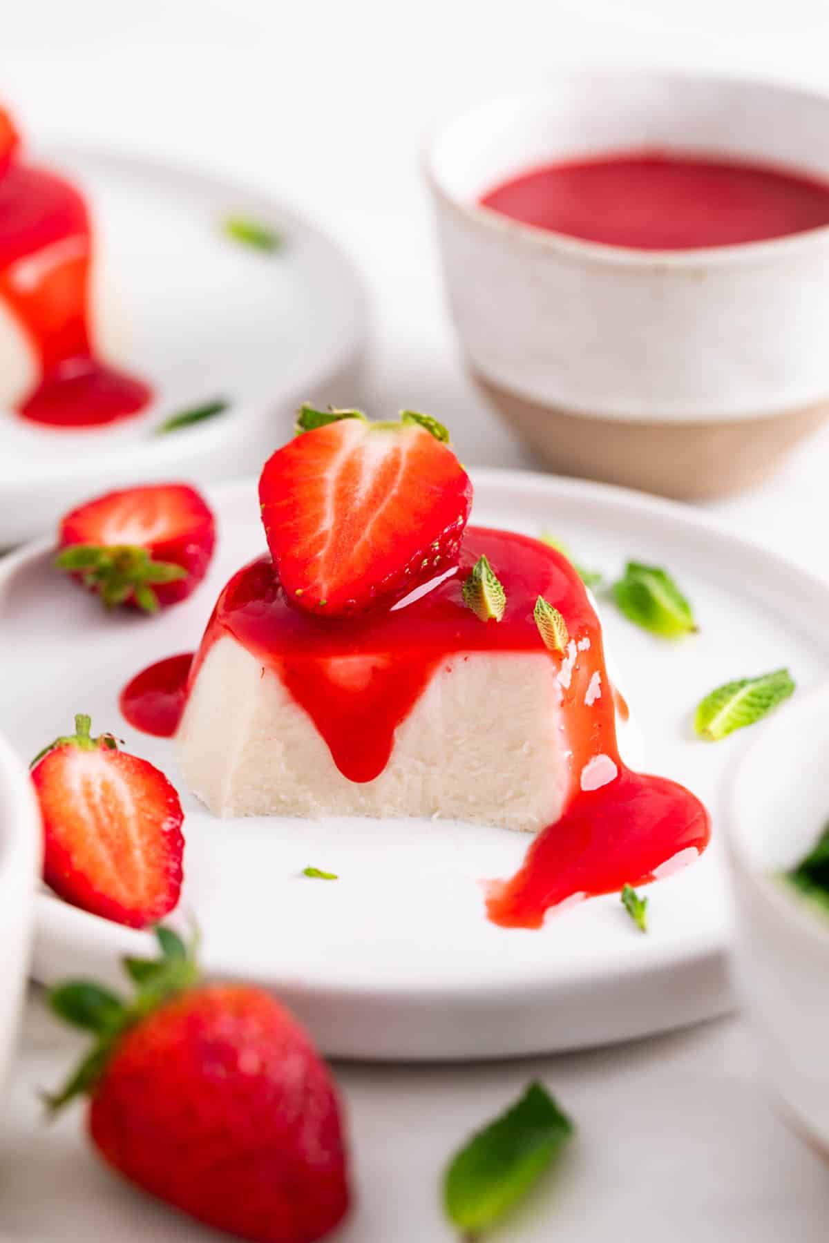 Vegan panna cotta on a plate, cut to reveal the inside, topped with strawberry coulis, a strawberry, and mint leaves.