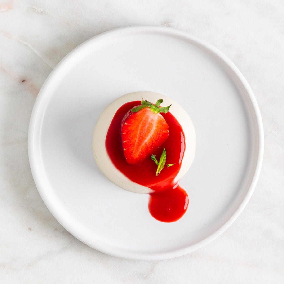 Vegan panna cotta on a plate with strawberry coulis and a strawberry on top.