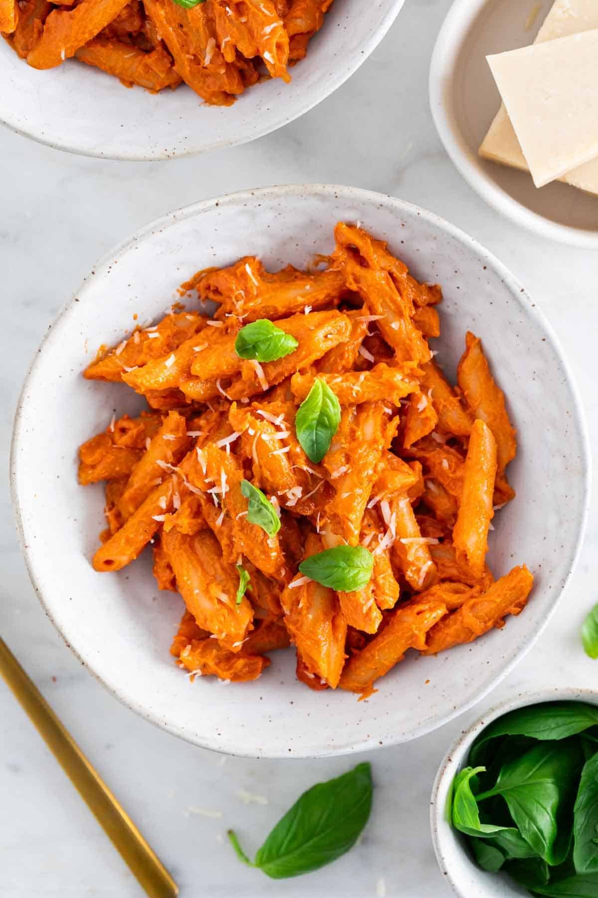 Vegan penne alla vodka in a bowl, with basil and vegan parmesan cheese surrounding it.