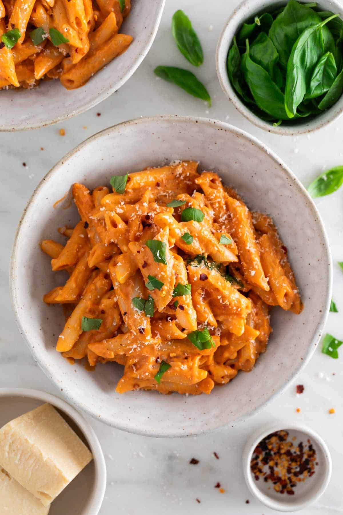 Penne with vegan pasta sauce in a bowl, topped with fresh basil and vegan Parmesan, with ingredients surrounding the bowl.