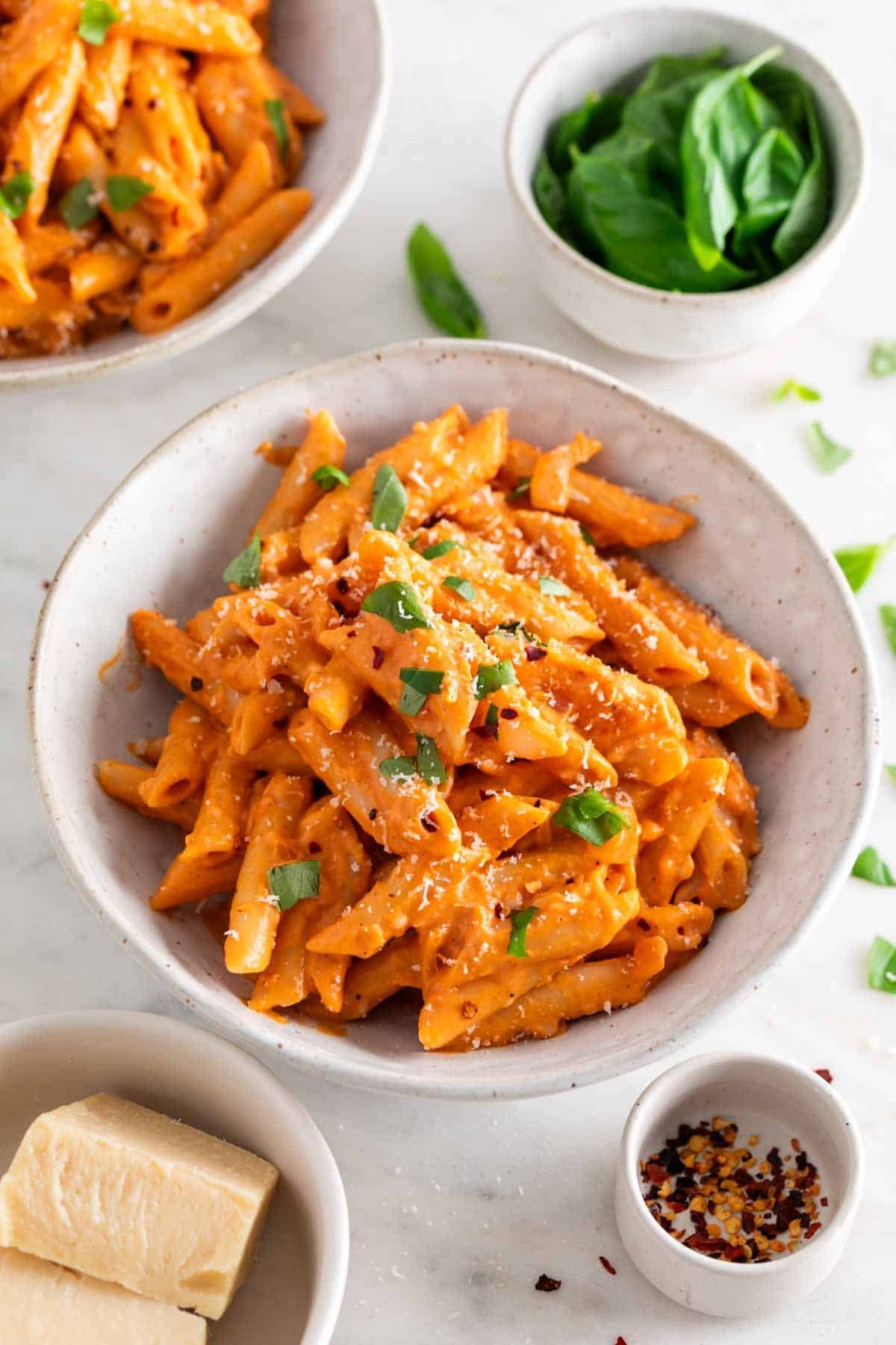 Penne with vegan pasta sauce in a bowl, garnished with fresh basil and vegan Parmesan, surrounded by ingredients.