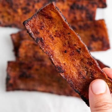 Tofu bacon slices on a white background with a finger picking up a slice.