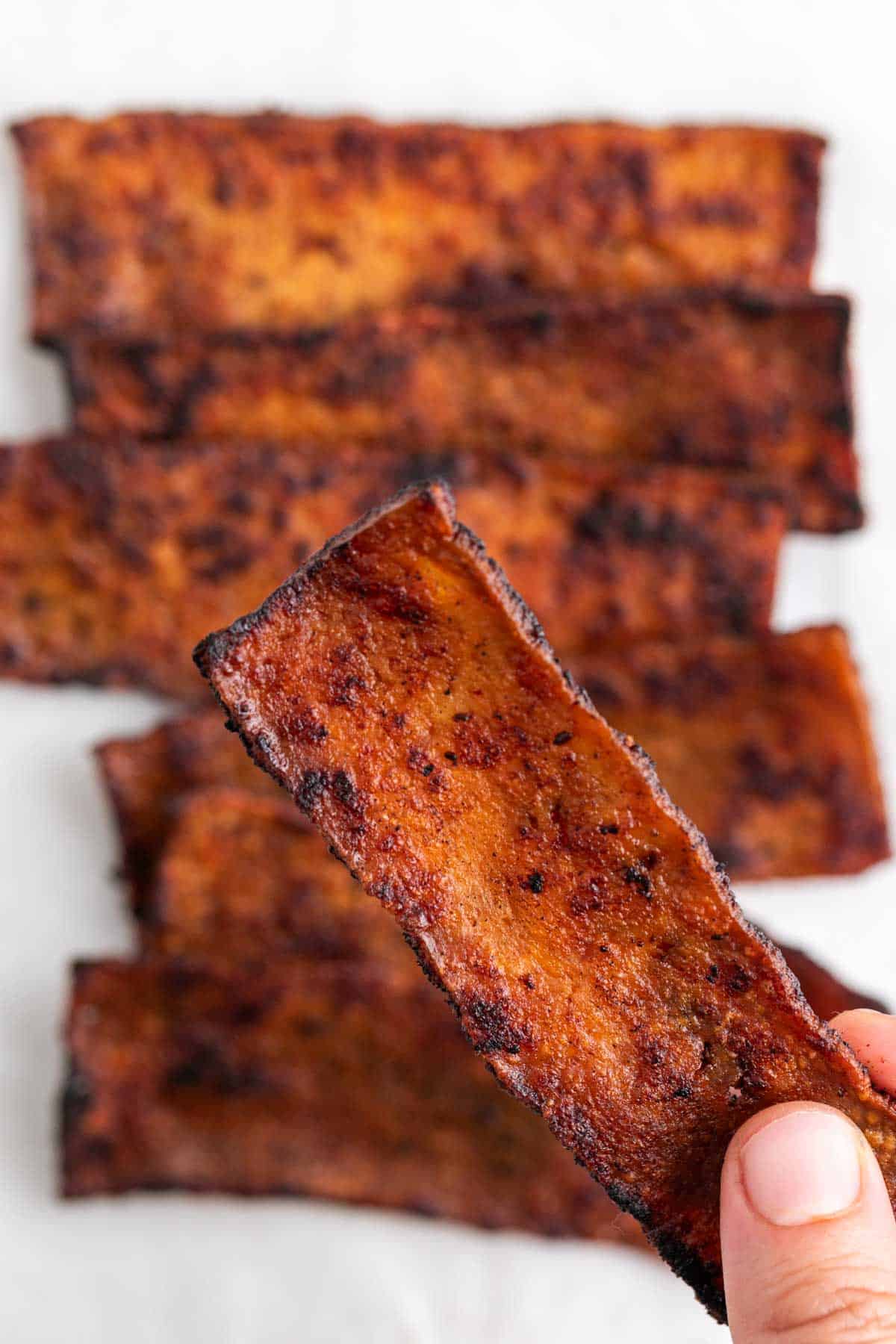 Tofu bacon slices on a white background with a finger picking up a slice.