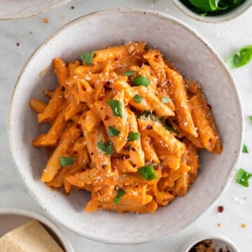 Penne with vegan pasta sauce in a bowl, topped with fresh basil and vegan Parmesan, with ingredients surrounding the bowl.