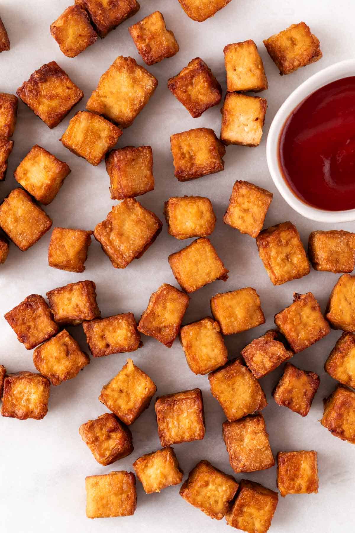 Crispy air fryer tofu on a white surface next to a small bowl of ketchup.