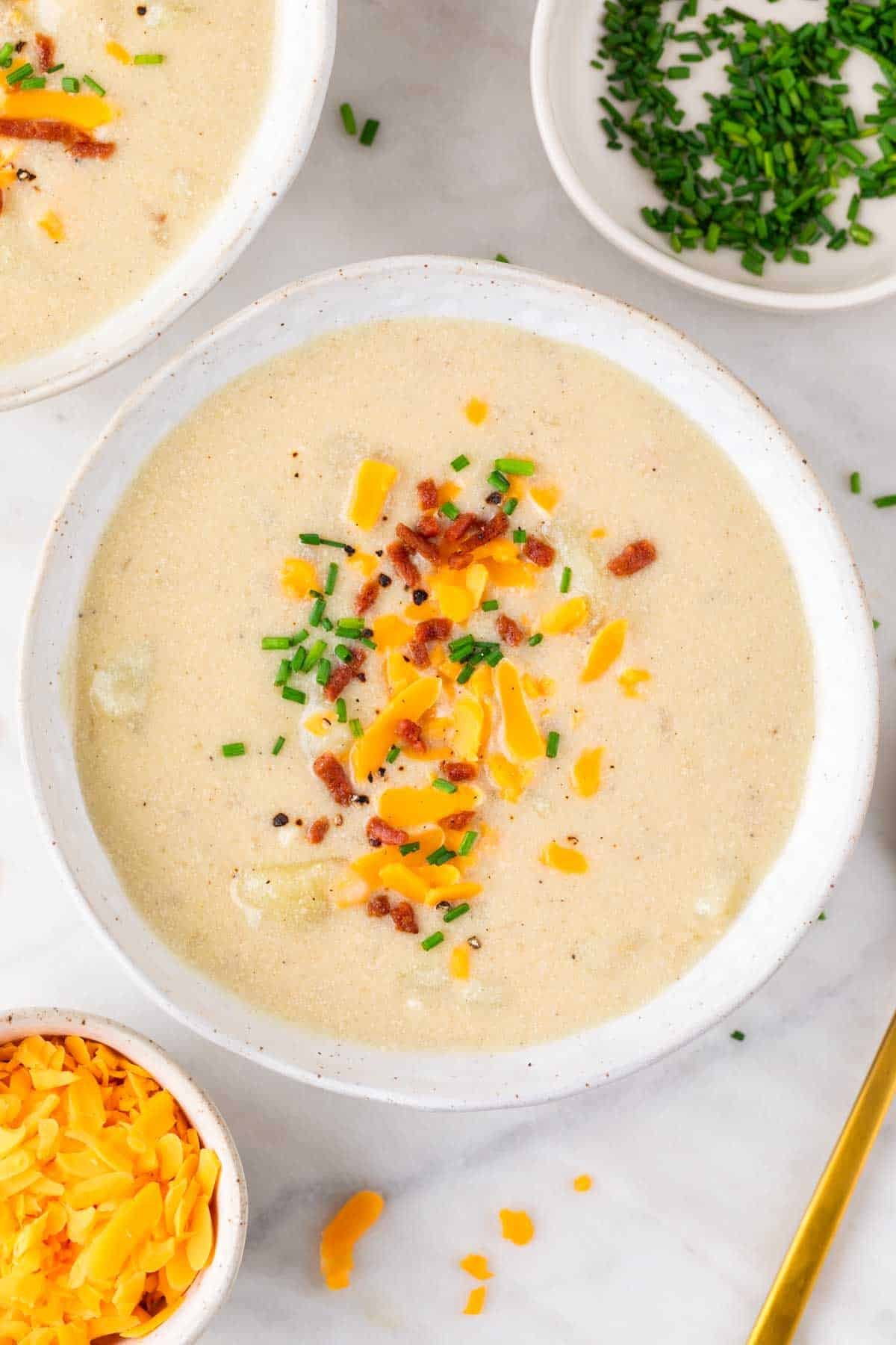 Two bowls of potato soup with potatoes, onions, and other ingredients around them.