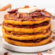 A tall stack of vegan pumpkin pancakes drizzled with maple syrup and topped with vegan butter and pecans.