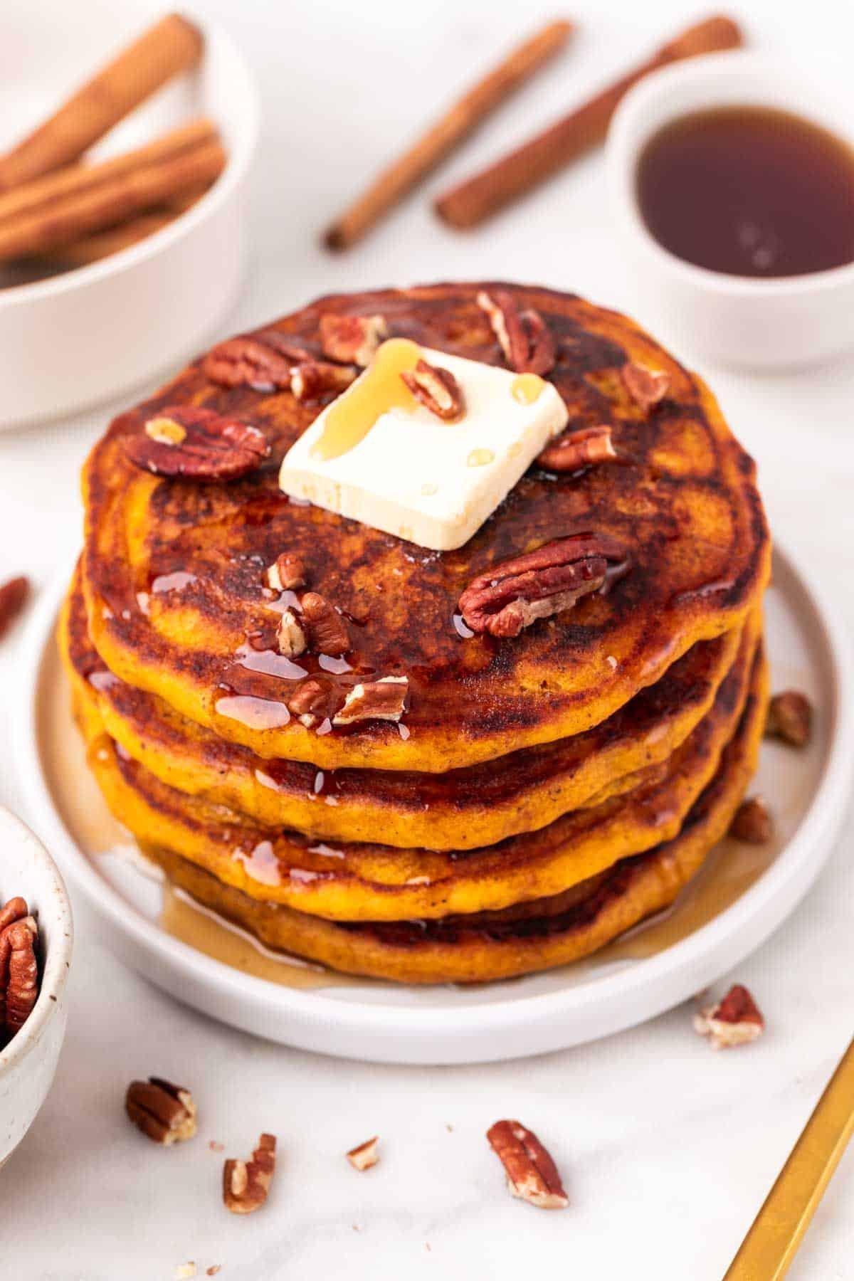 Pumpkin pancake stack topped with syrup, butter, and pecans, with ingredients around.
