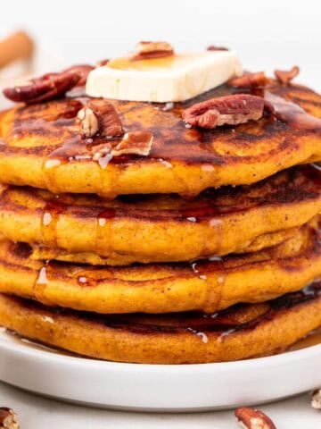 A tall stack of vegan pumpkin pancakes drizzled with maple syrup and topped with vegan butter and pecans.