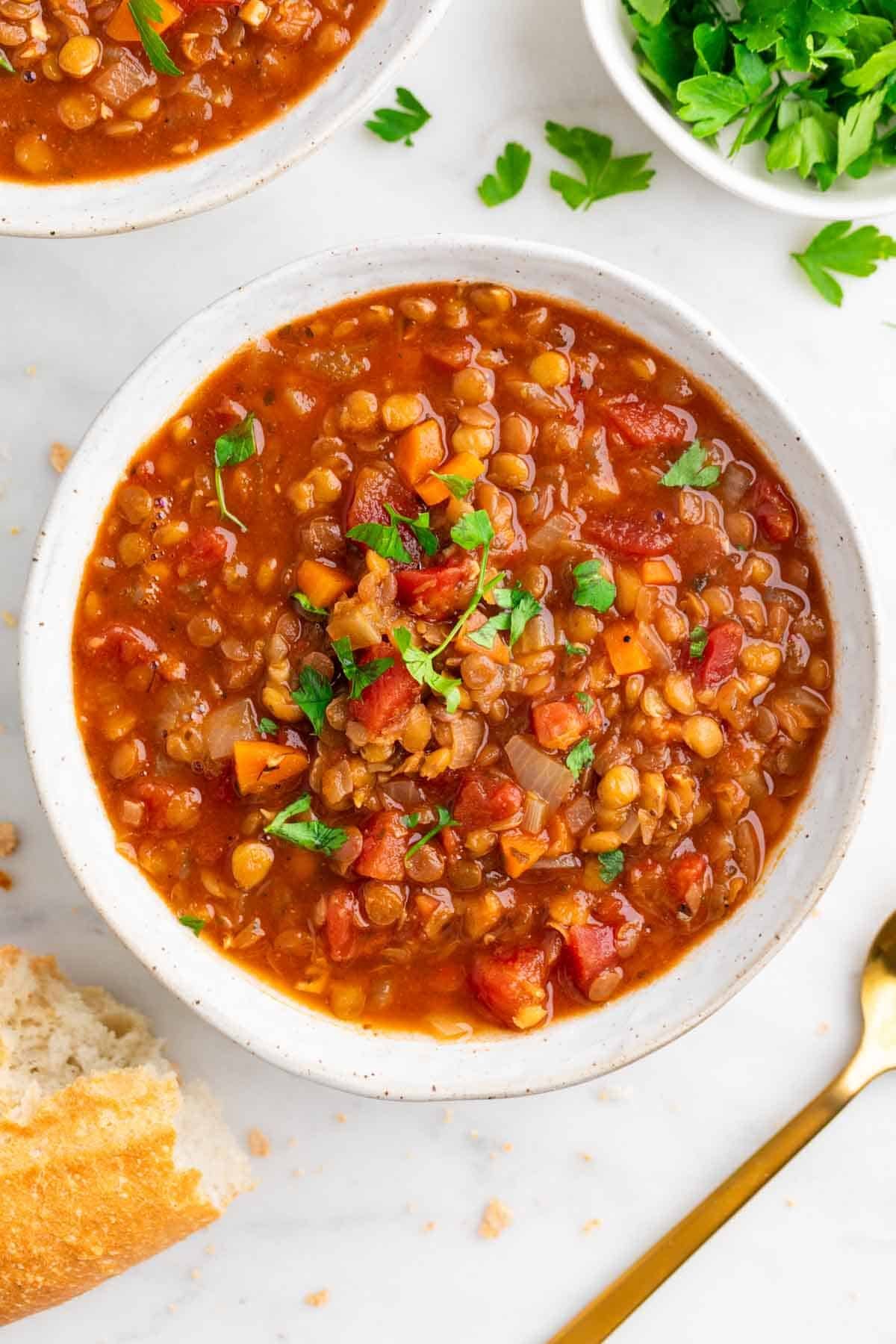 Bowl of vegan lentil soup with a spoon, crusty bread, and fresh parsley on the side.