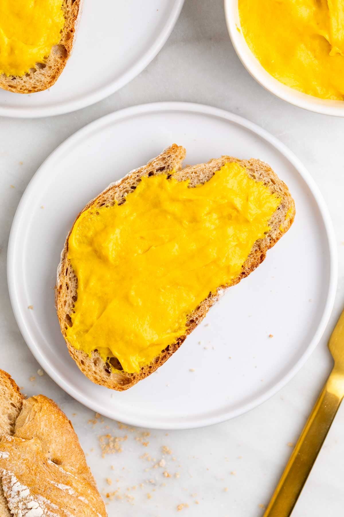 Slice of bread with vegan cheddar cheese spread on a plate with extra bread and a knife.
