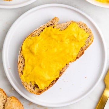 Slice of bread spread with vegan cheddar cheese on a plate with extra slices and a knife.
