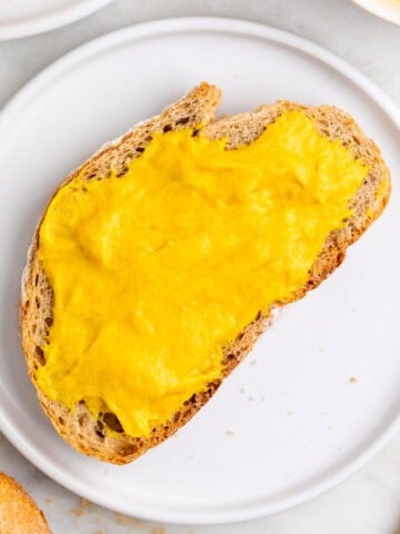 Slice of bread spread with vegan cheddar cheese on a plate with extra slices and a knife.