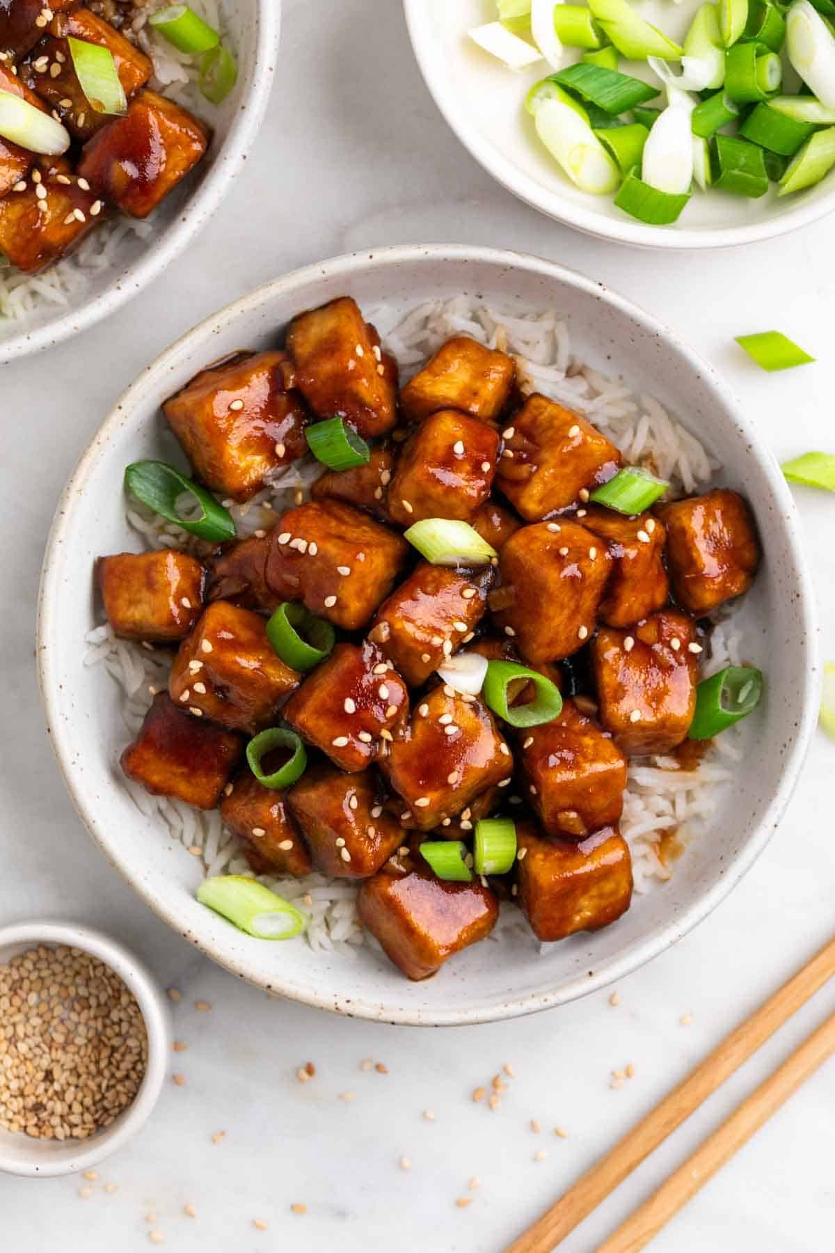 Bowl of Mongolian tofu over rice topped with sesame seeds and scallions.