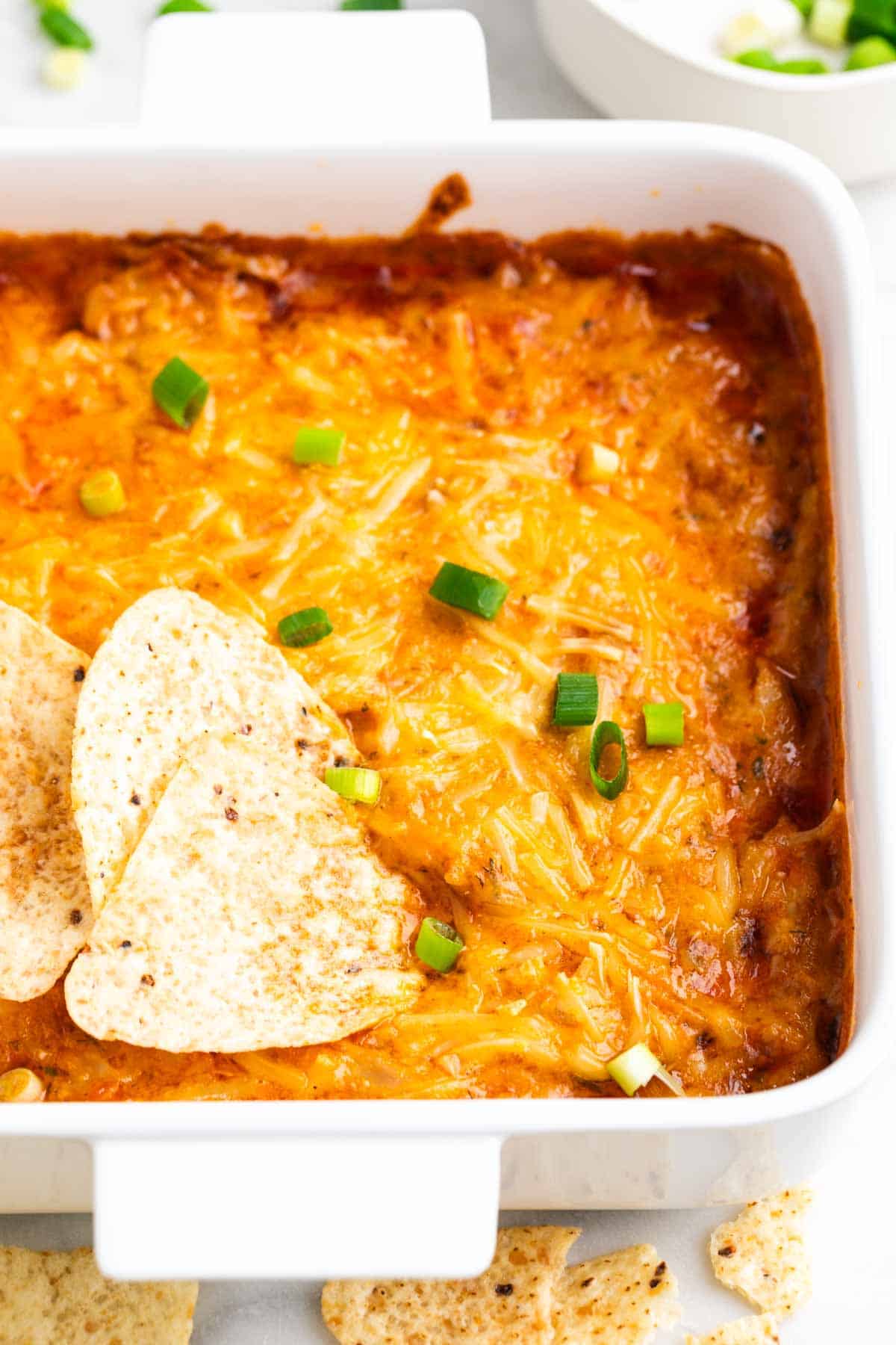 Baked vegan buffalo chicken dip topped with scallions, with tortilla chips on the side.