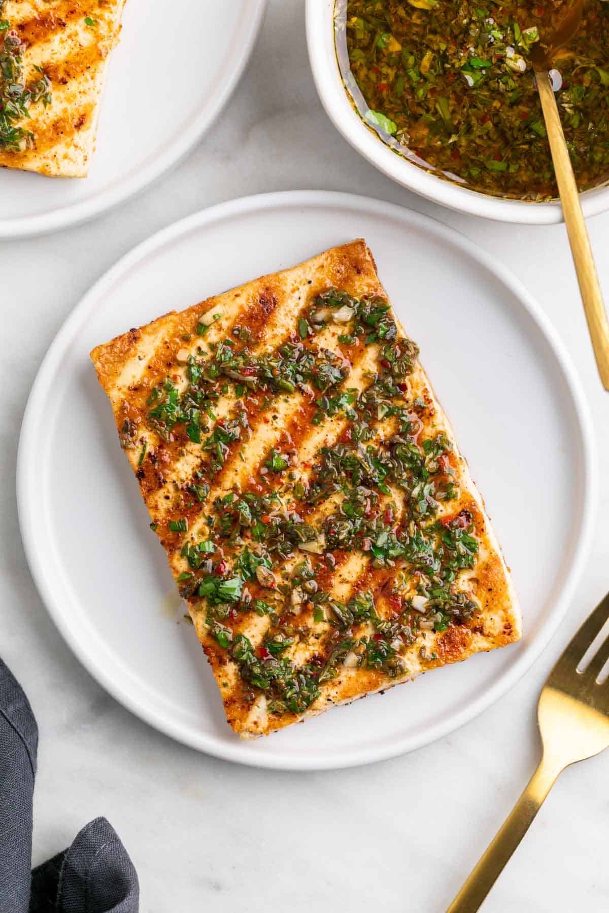 Two plates with grilled tofu steaks topped with chimichurri, with a fork and extra chimichurri on the side.