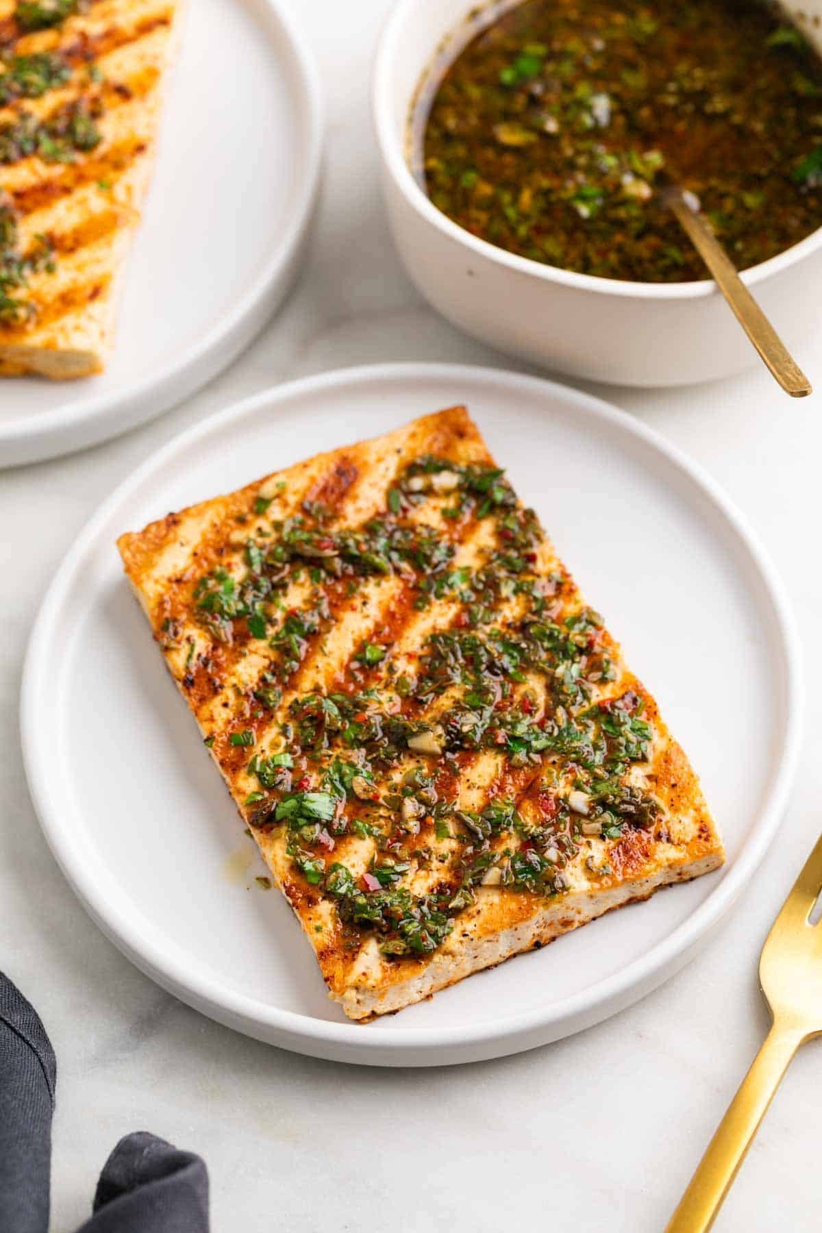 Two plates of grilled tofu with chimichurri, served with a fork and extra sauce.