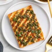 Two plates of grilled tofu steaks topped with chimichurri, with a fork and extra chimichurri around.