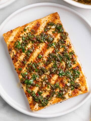 Two plates of grilled tofu steaks topped with chimichurri, with a fork and extra chimichurri around.
