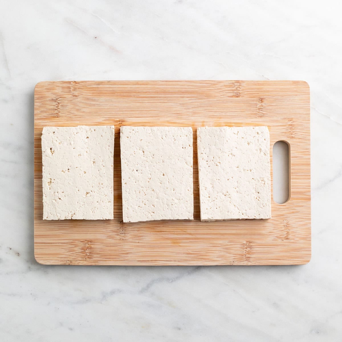 Block of tofu cut into three thick slabs on a cutting board.