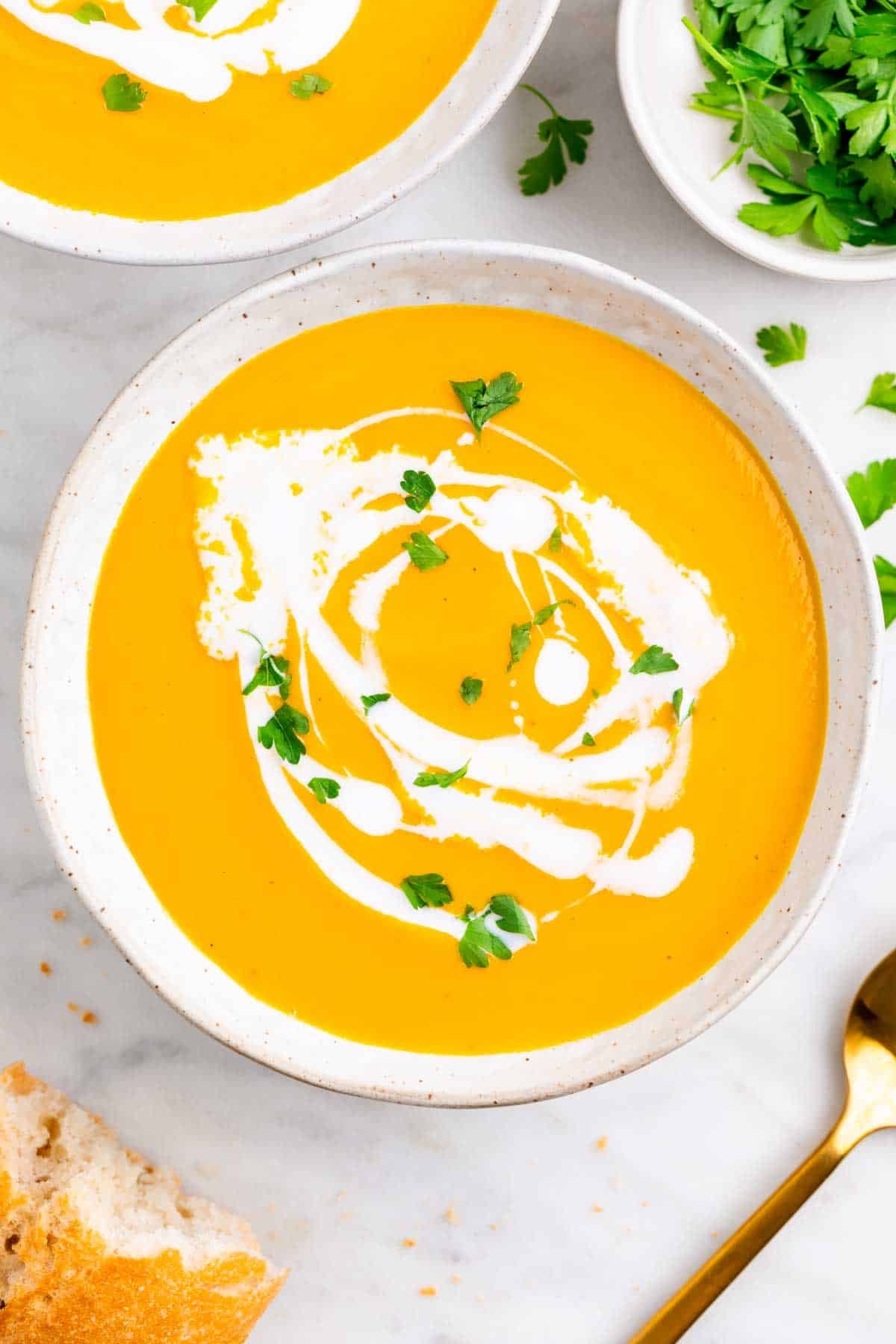 Two bowls of vegan carrot soup with coconut milk, parsley, bread, and a spoon on the side