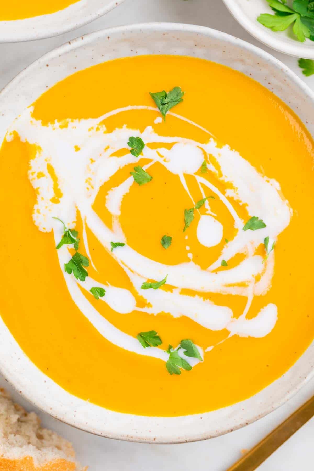 Bowls of vegan carrot soup with coconut milk and parsley, with bread and a spoon nearby