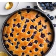 Vegan lemon blueberry cake in a pan with fresh lemon, blueberries, and powdered sugar nearby