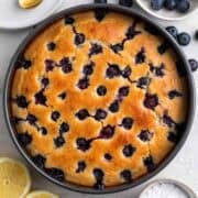 Vegan lemon blueberry cake in a pan with lemon halves, powdered sugar, and blueberries