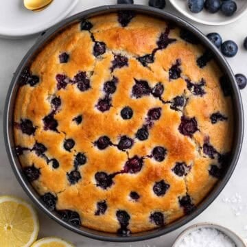 Vegan lemon blueberry cake in a pan with lemon halves, powdered sugar, and blueberries