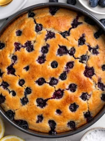 Vegan lemon blueberry cake in a pan with lemon halves, powdered sugar, and blueberries