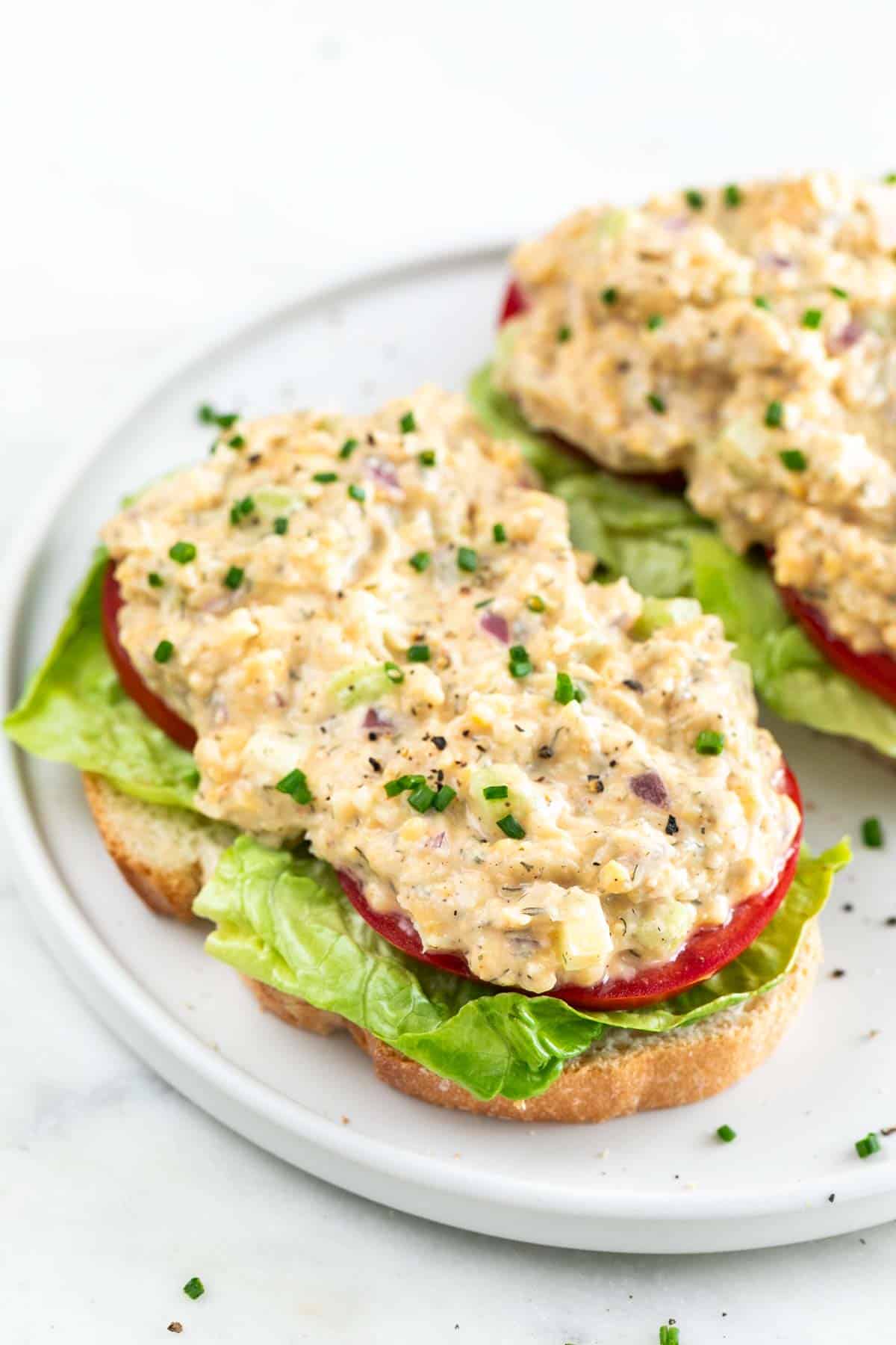 Two open-faced vegan chicken salad sandwiches with lettuce and tomato on a white plate