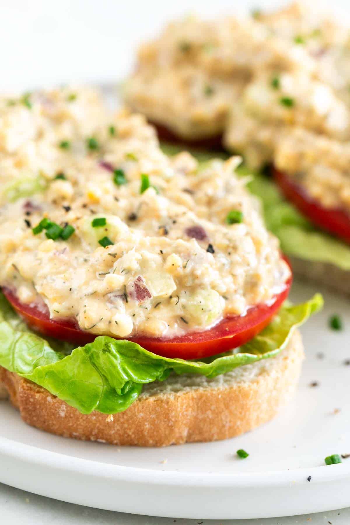 Two slices of toast topped with lettuce, tomato, vegan chicken salad, chives, and black pepper