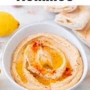 Homemade hummus in a bowl with paprika, olive oil, pita bread, lemons, and garlic