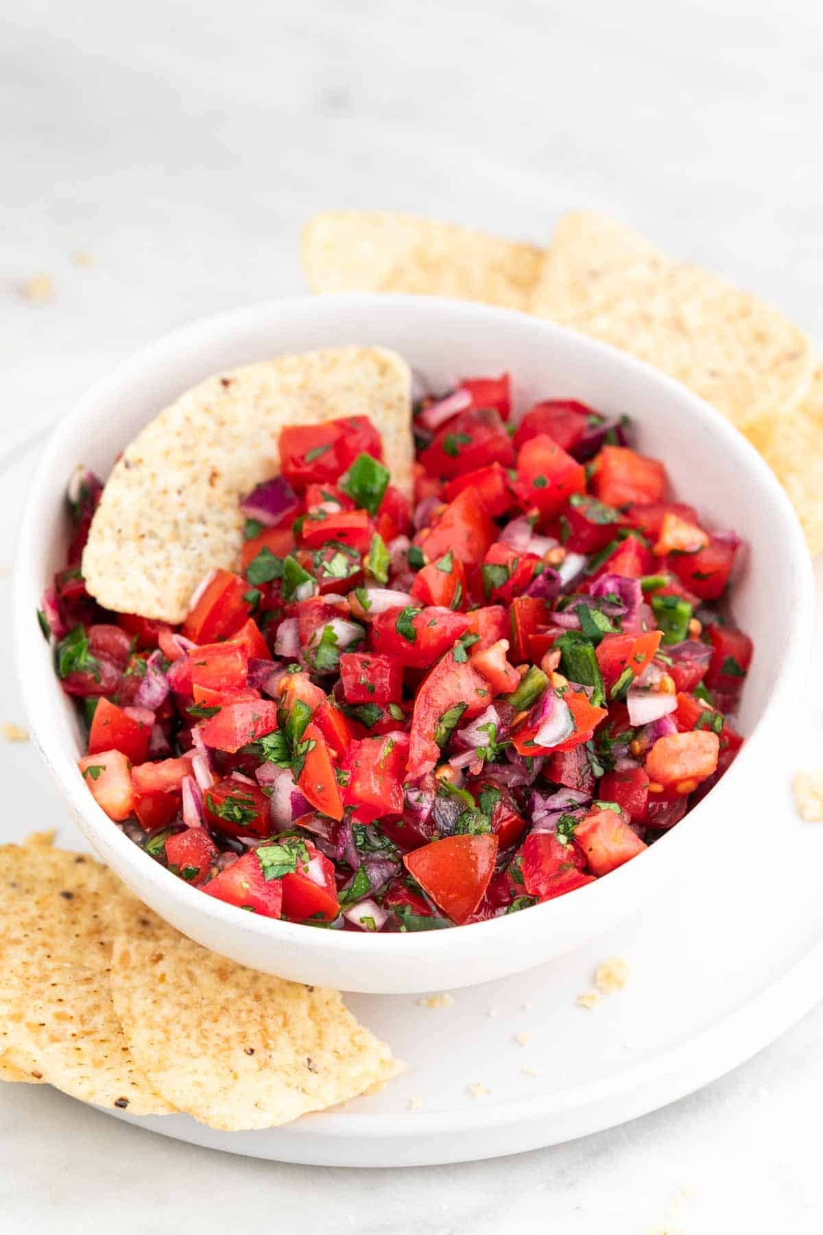 Pico de gallo in a white bowl with tortilla chips inside and scattered around