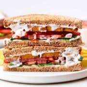 Vegan BLT sandwich halves stacked on a white plate with potato chips on the side