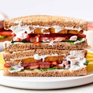 Vegan BLT sandwich halves stacked on a white plate with potato chips on the side