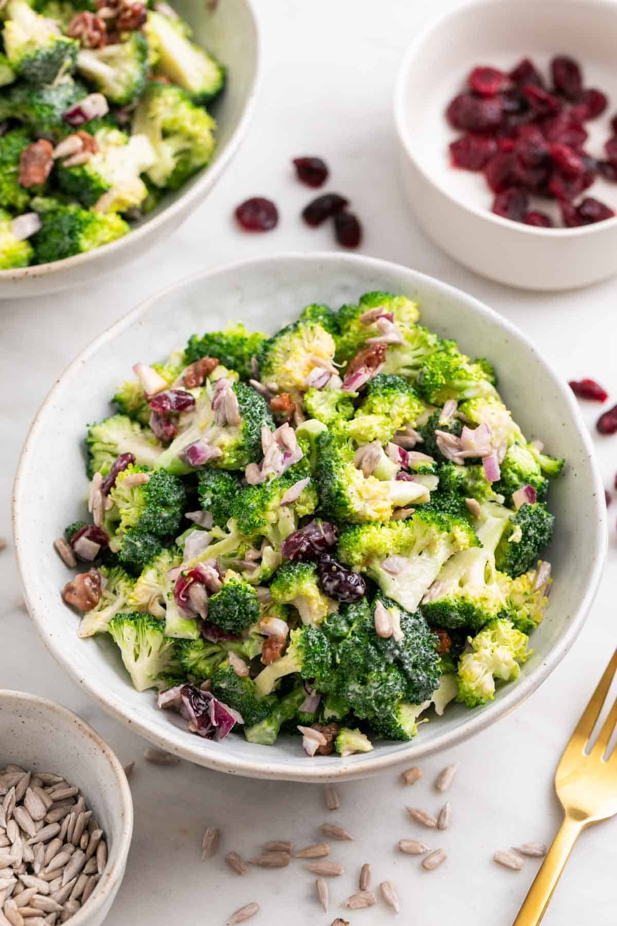 Vegan broccoli salad served in two bowls with dried cranberries nearby