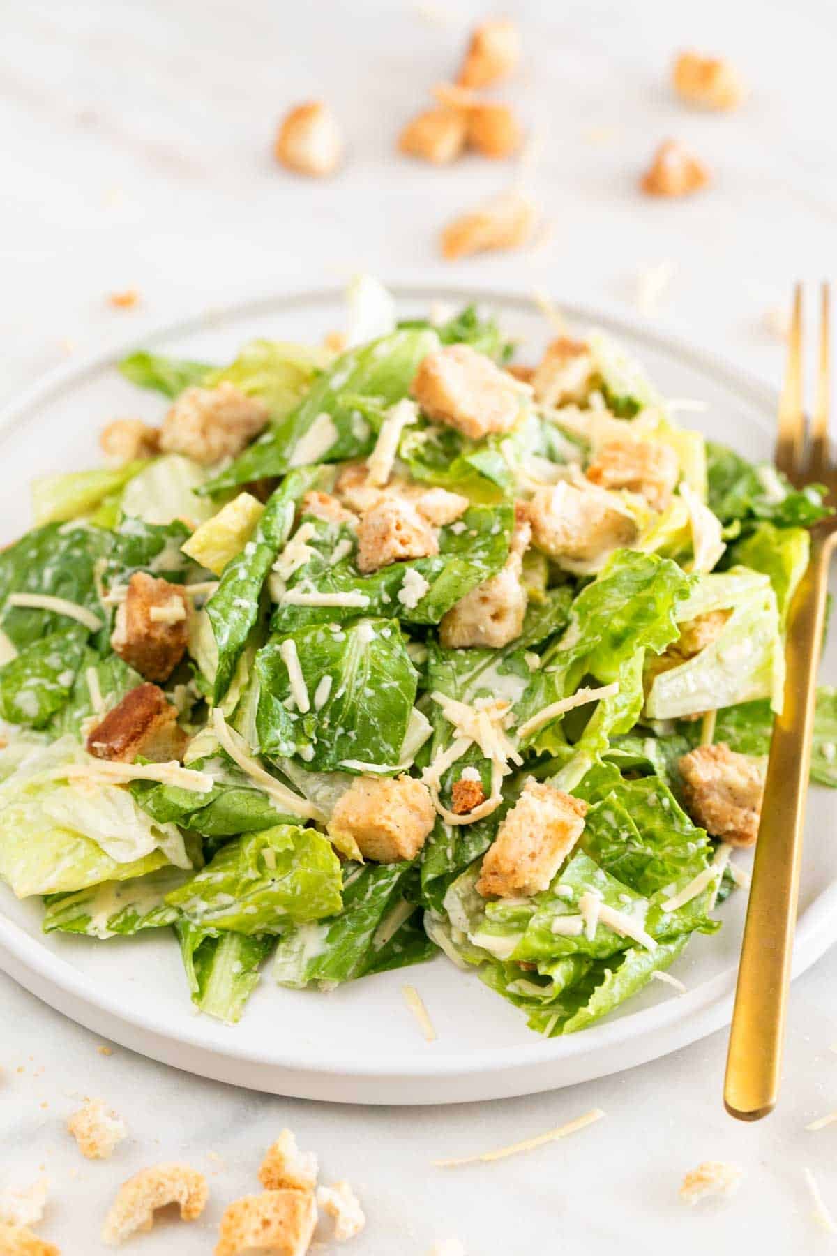 Plate of vegan Caesar salad with a fork, croutons, and extra vegan Parmesan on a marble surface