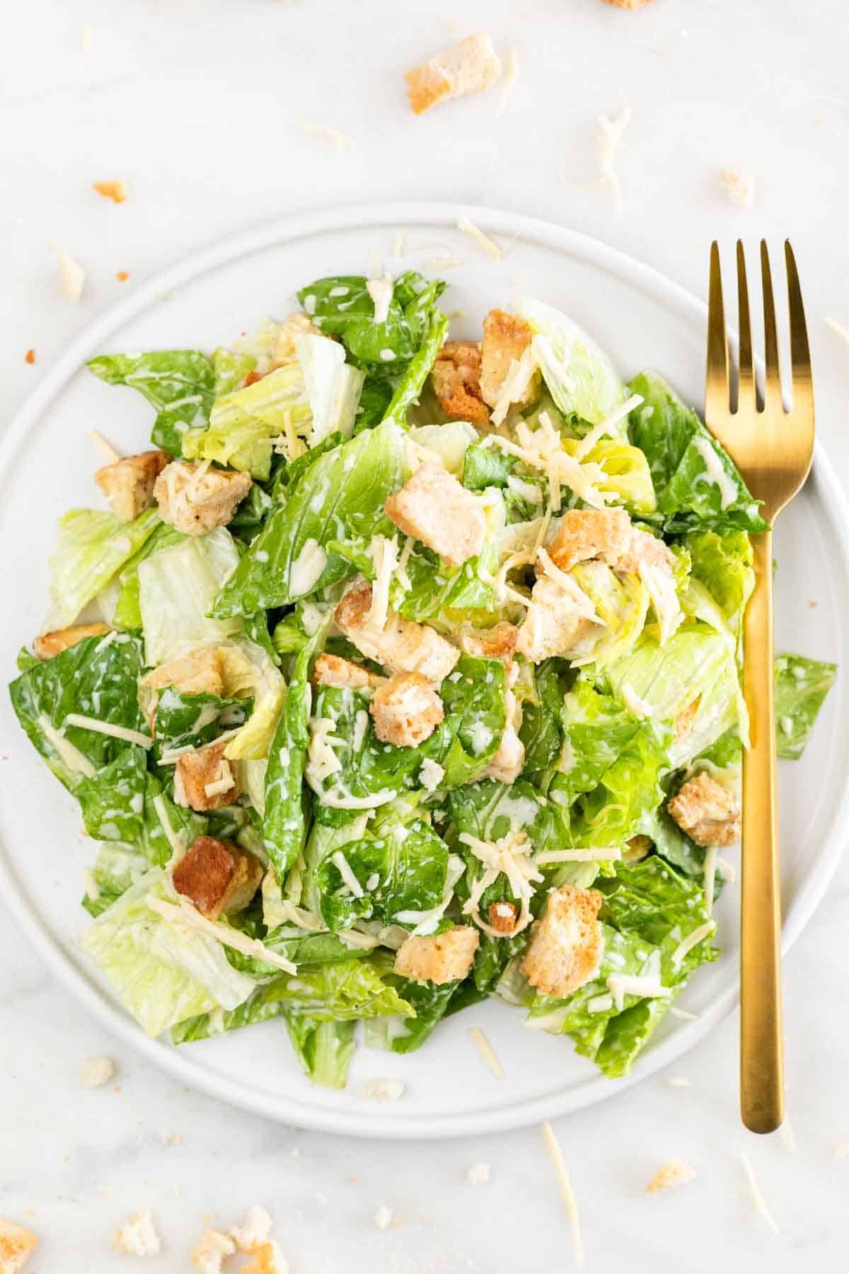 Plate of vegan Caesar salad with a fork and extra vegan Parmesan on a marble surface